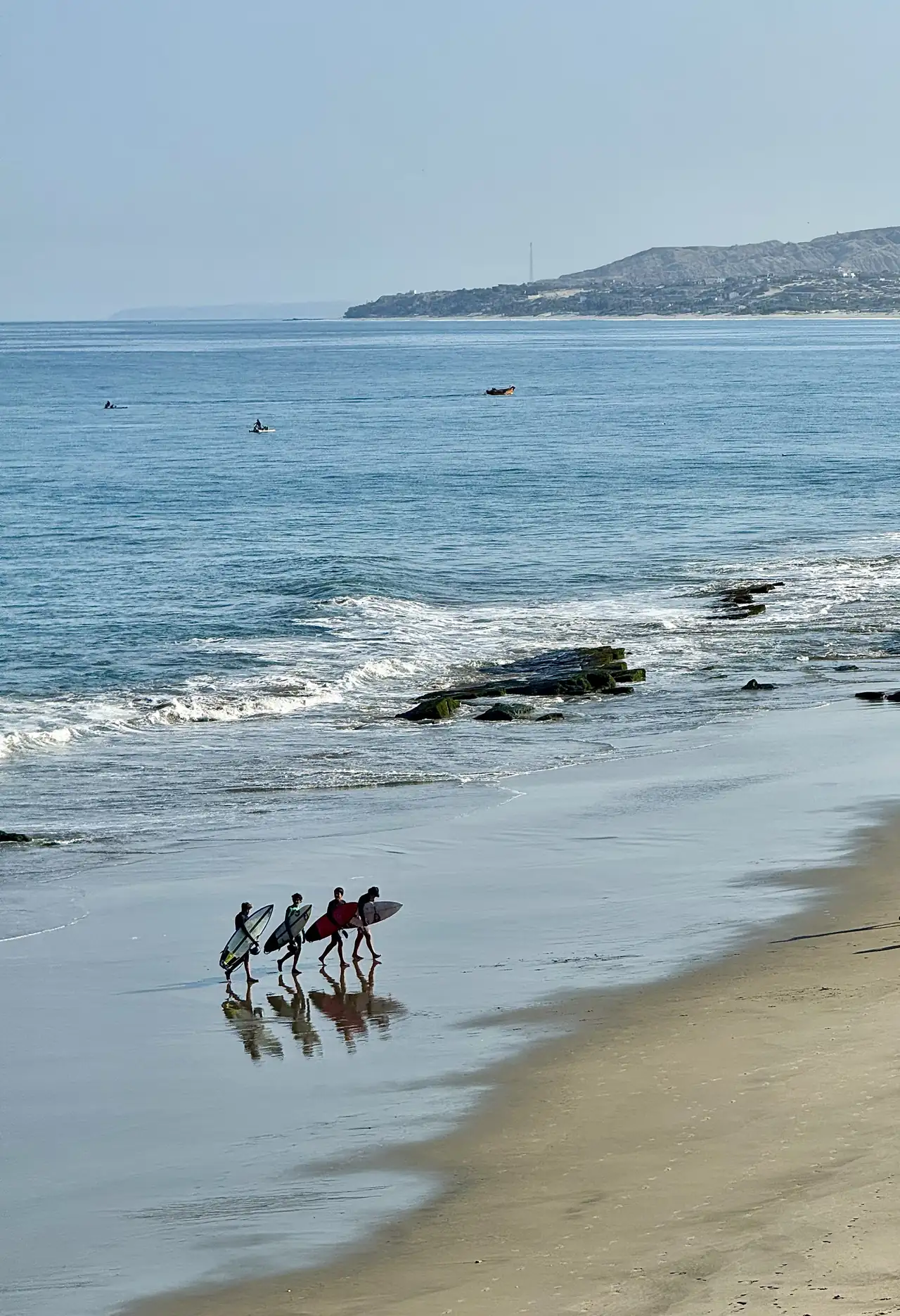 Surfistas con sus tablas en la playa, con botes y la costa al fondo.