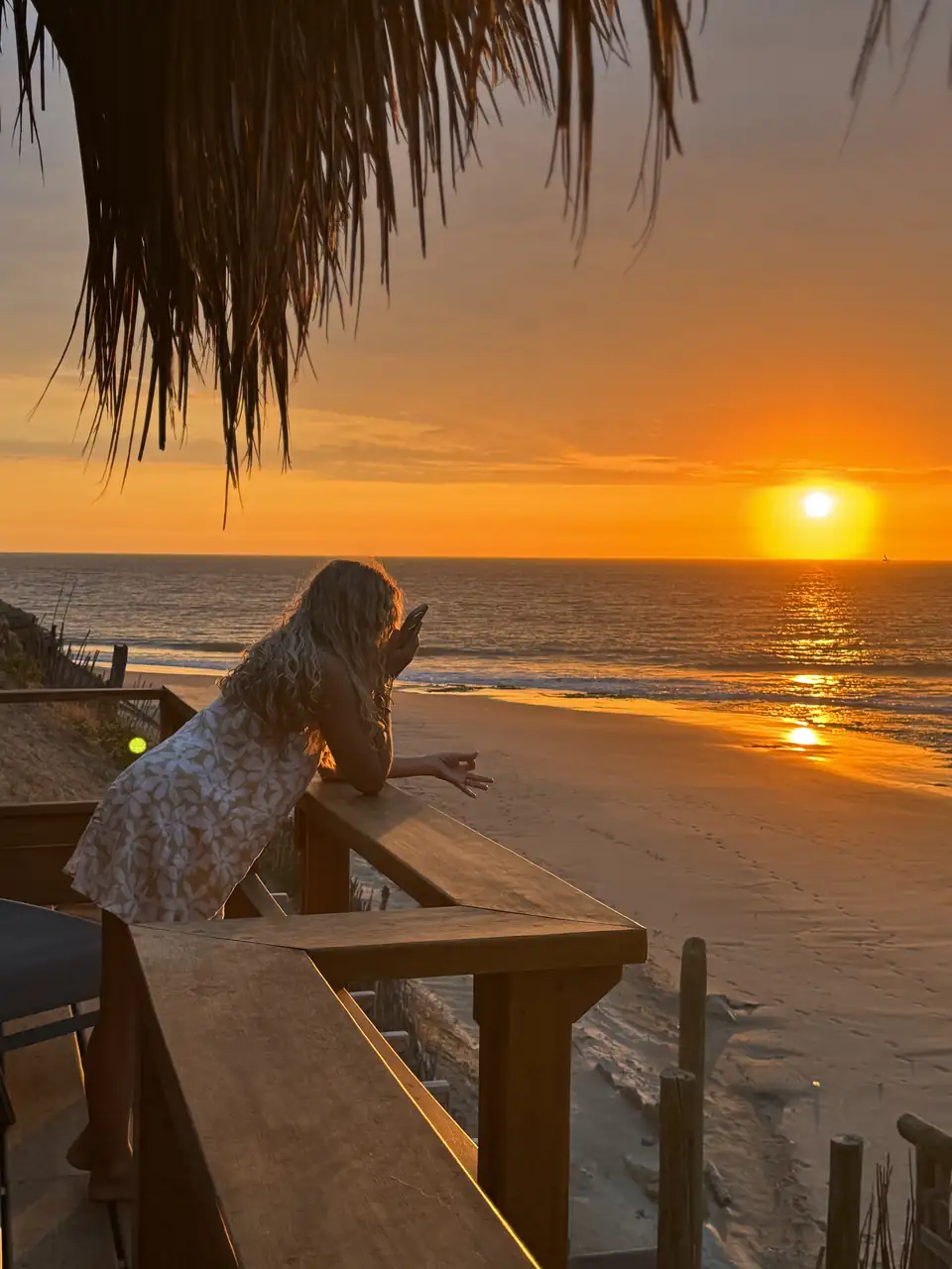 Una persona en una terraza de madera frente a la playa y al océano al atardecer.